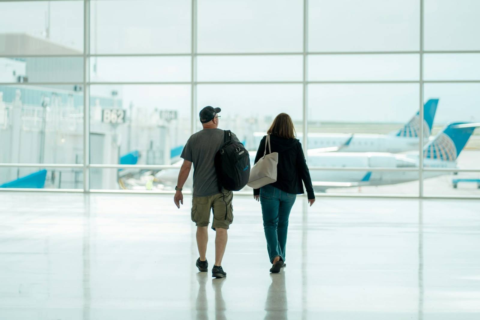 Two travellers with backpacks walking through a departure hall, planes behind the glass