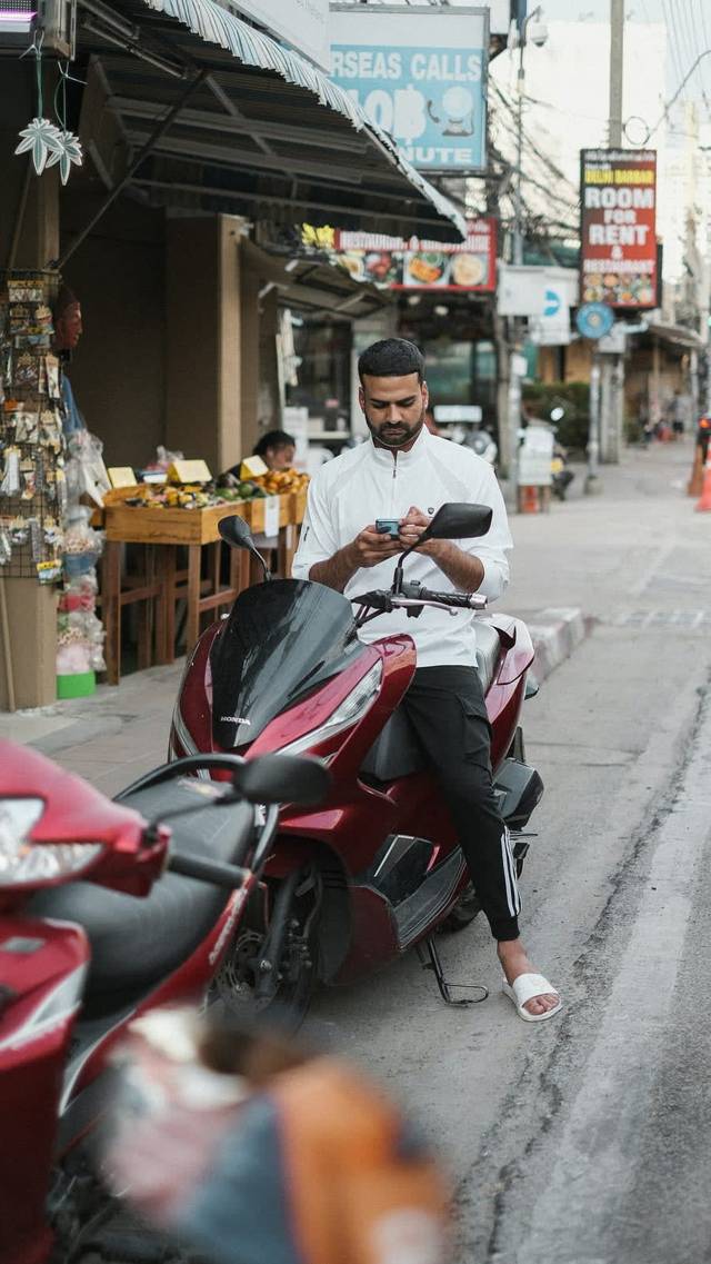 A man on a parked scooter looking at his phone, with a sign advertising international calls behind him